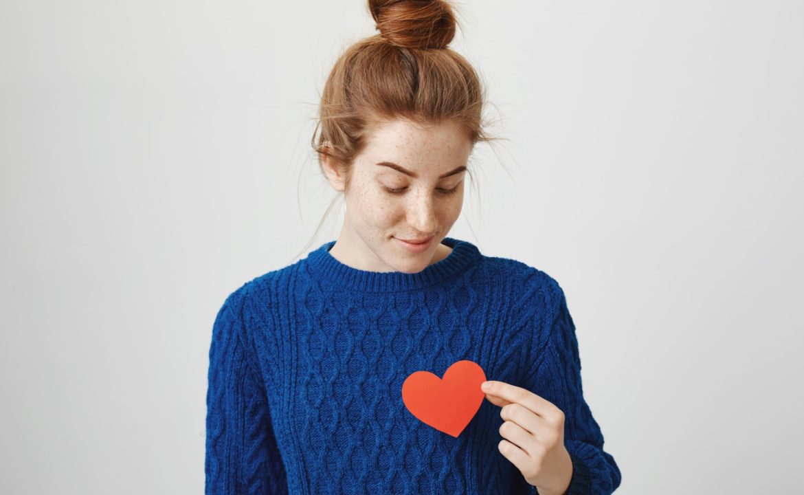 Lonely soul in big city. Indoor shot of attractive redhead woman with bun in cozy winter sweater holding paper heart near chest and looking with dreamy expression, being hopeless romantic