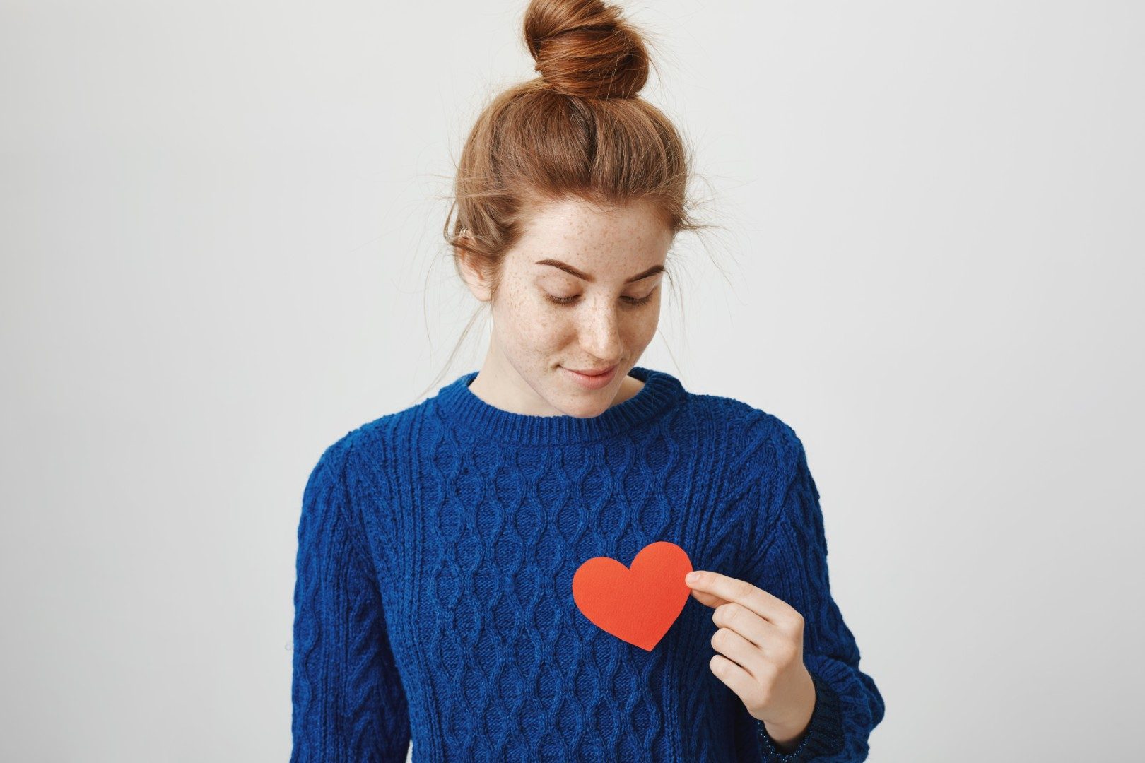 Lonely soul in big city. Indoor shot of attractive redhead woman with bun in cozy winter sweater holding paper heart near chest and looking with dreamy expression, being hopeless romantic