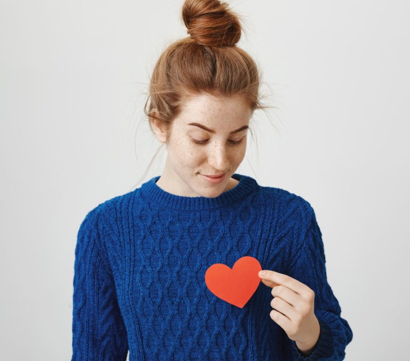 Lonely soul in big city. Indoor shot of attractive redhead woman with bun in cozy winter sweater holding paper heart near chest and looking with dreamy expression, being hopeless romantic