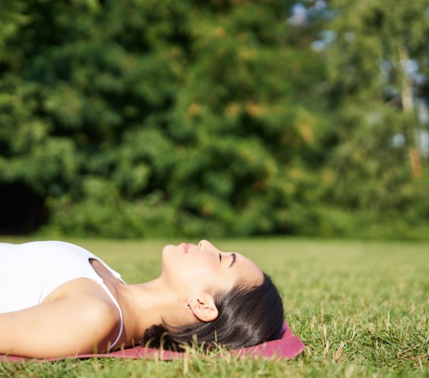 Young fitness girl lying on sport mat on lawn, breathing and meditating in park in sportswear