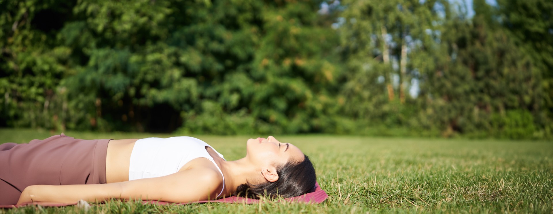 Young fitness girl lying on sport mat on lawn, breathing and meditating in park in sportswear
